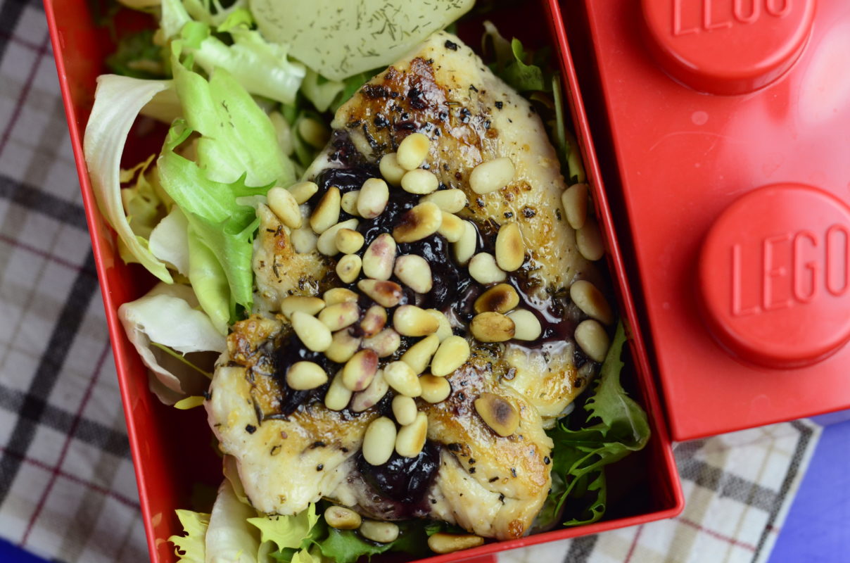 Kurczak z sosem porzeczkowym posypany orzeszkami pini, ułożony na sałacie. Kurczak w pojemniku typu lunchbox.