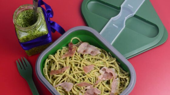 Spaghetti z pesto z rukolą i szynką parmeńską w zielonym lunchboxie. Obok widelec i słoiczek z pesto przewiązany wstążką niebieską.