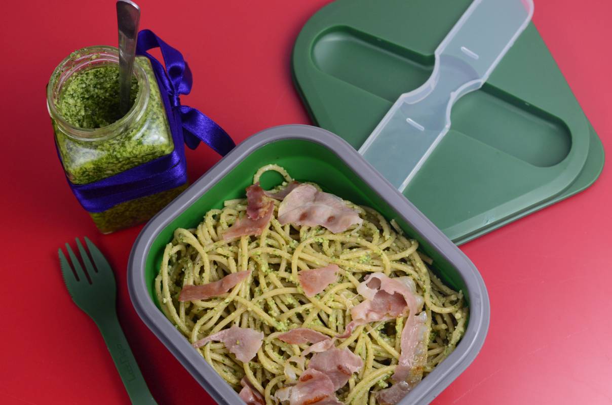 Spaghetti z pesto z rukolą i szynką parmeńską w zielonym lunchboxie. Obok widelec i słoiczek z pesto przewiązany wstążką niebieską.