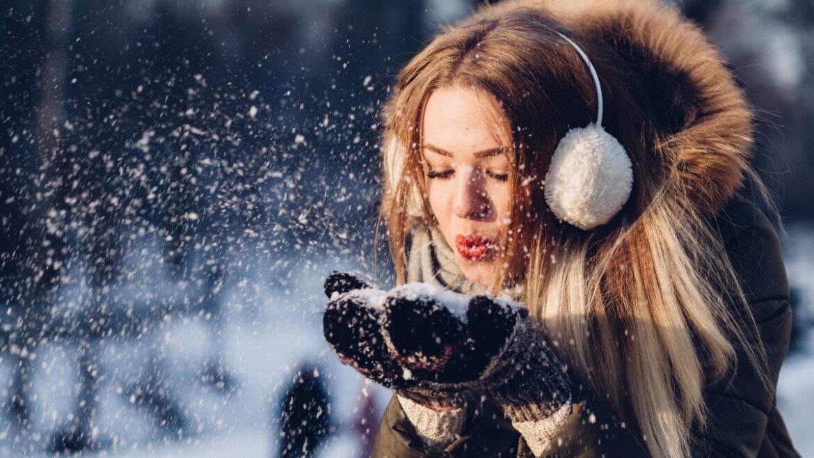Młoda kobiety dmucha w śnieg zebrany w dłoniach. Kobieta w ciepłej kurtce, nausznikach i rękawiczkach.