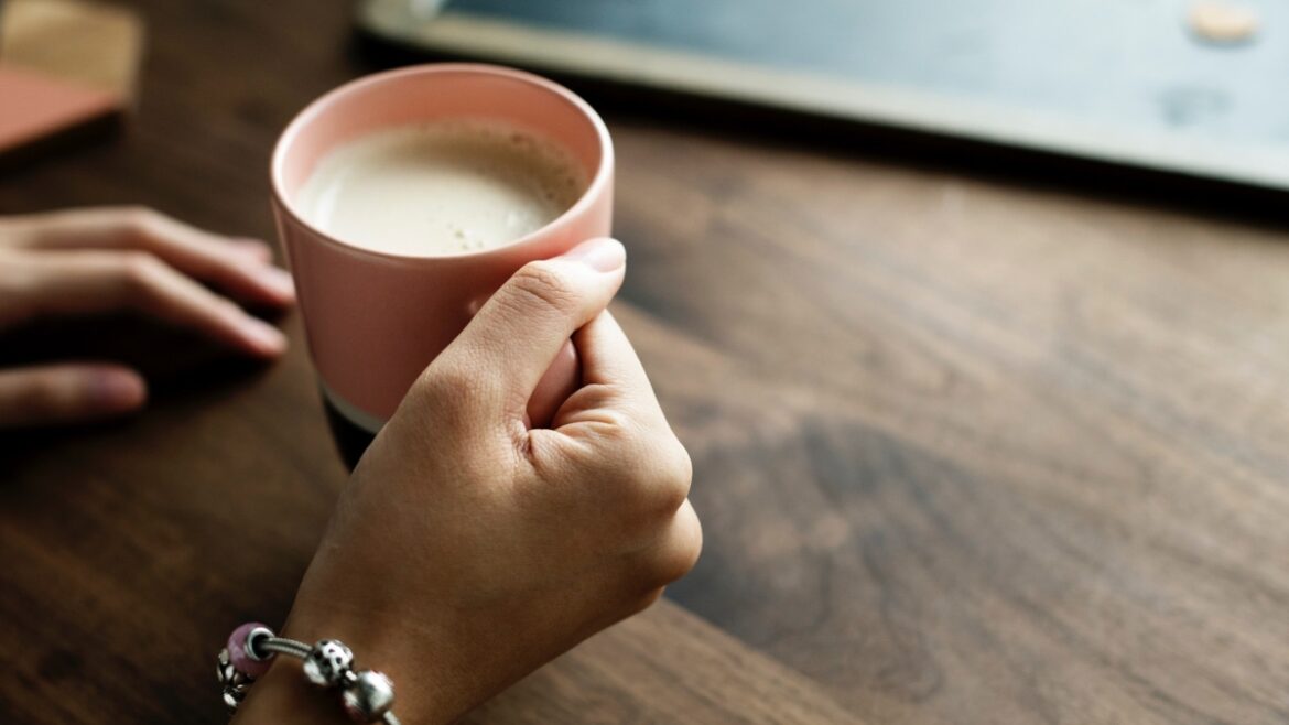 Kobieca dłoń trzyma kubek z kawą z mlekiem bez laktozy. Dolegliwością kobiety jest nietolerancja laktozy.